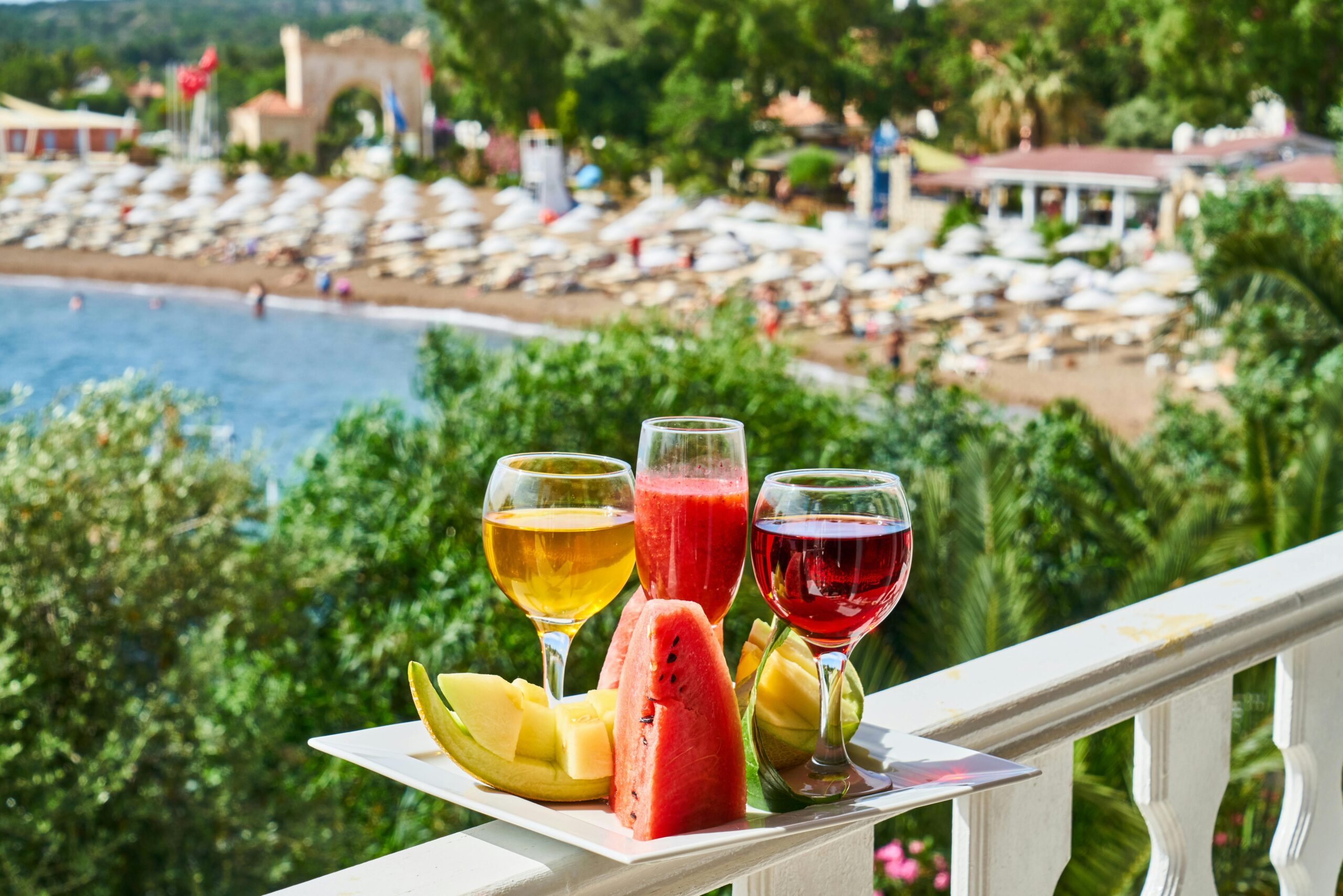 Tropical drinks and fresh fruits on a balcony overlooking a vibrant beach resort, perfect for a summer vacation setting.